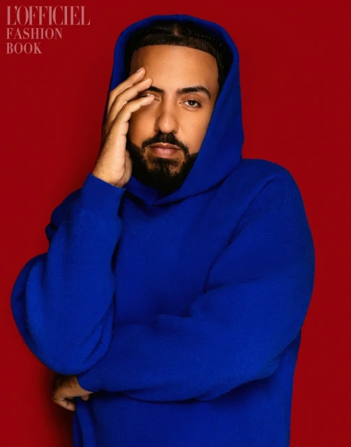 Portrait of a Black male celebrity wearing a blue hooded sweatshirt against a red studio backdrop for L'Officiel Fashion Book.