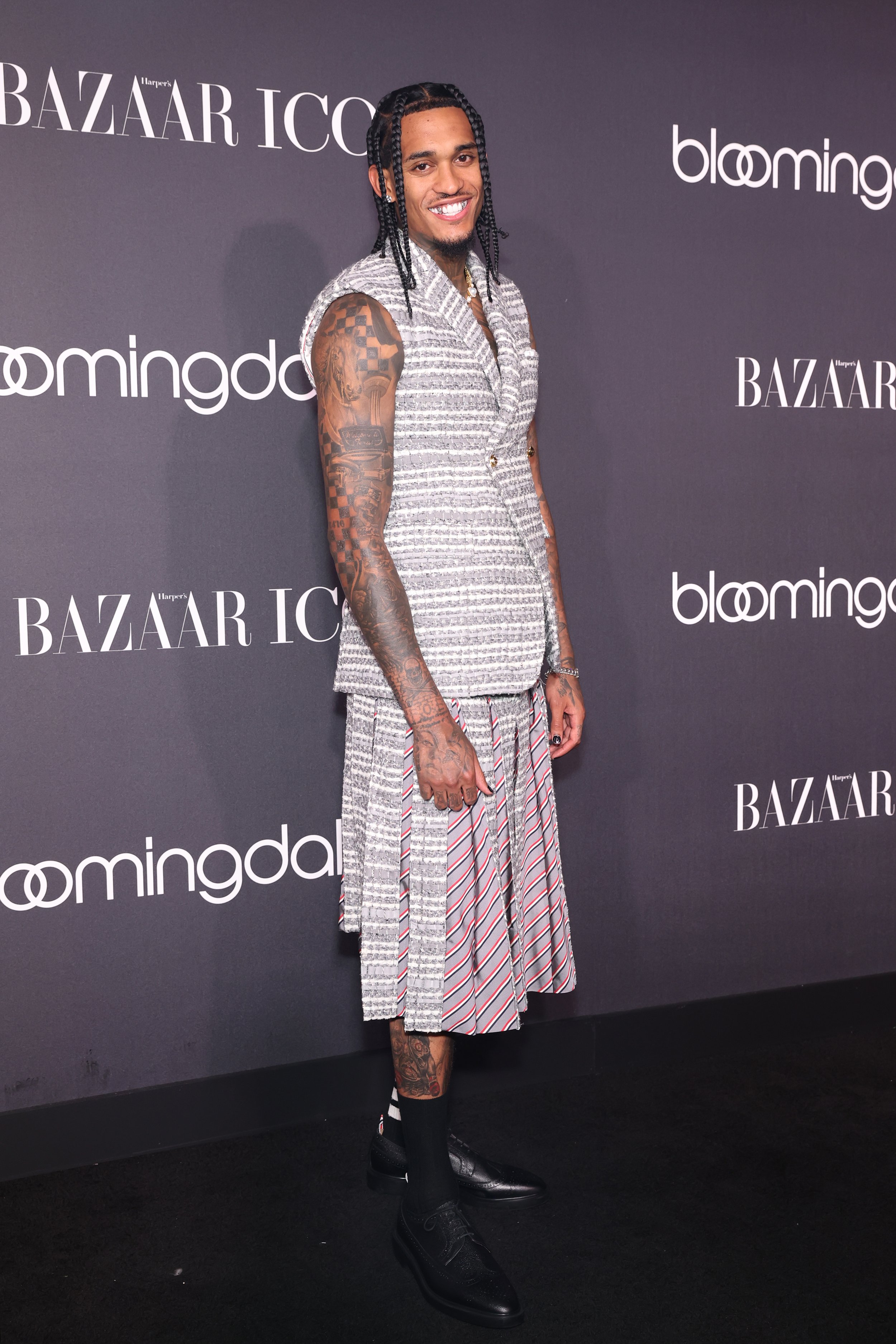 A smiling adult male with dark hair, wearing a sleeveless grey suit jacket and pleated skirt, posing in front of event branding.
