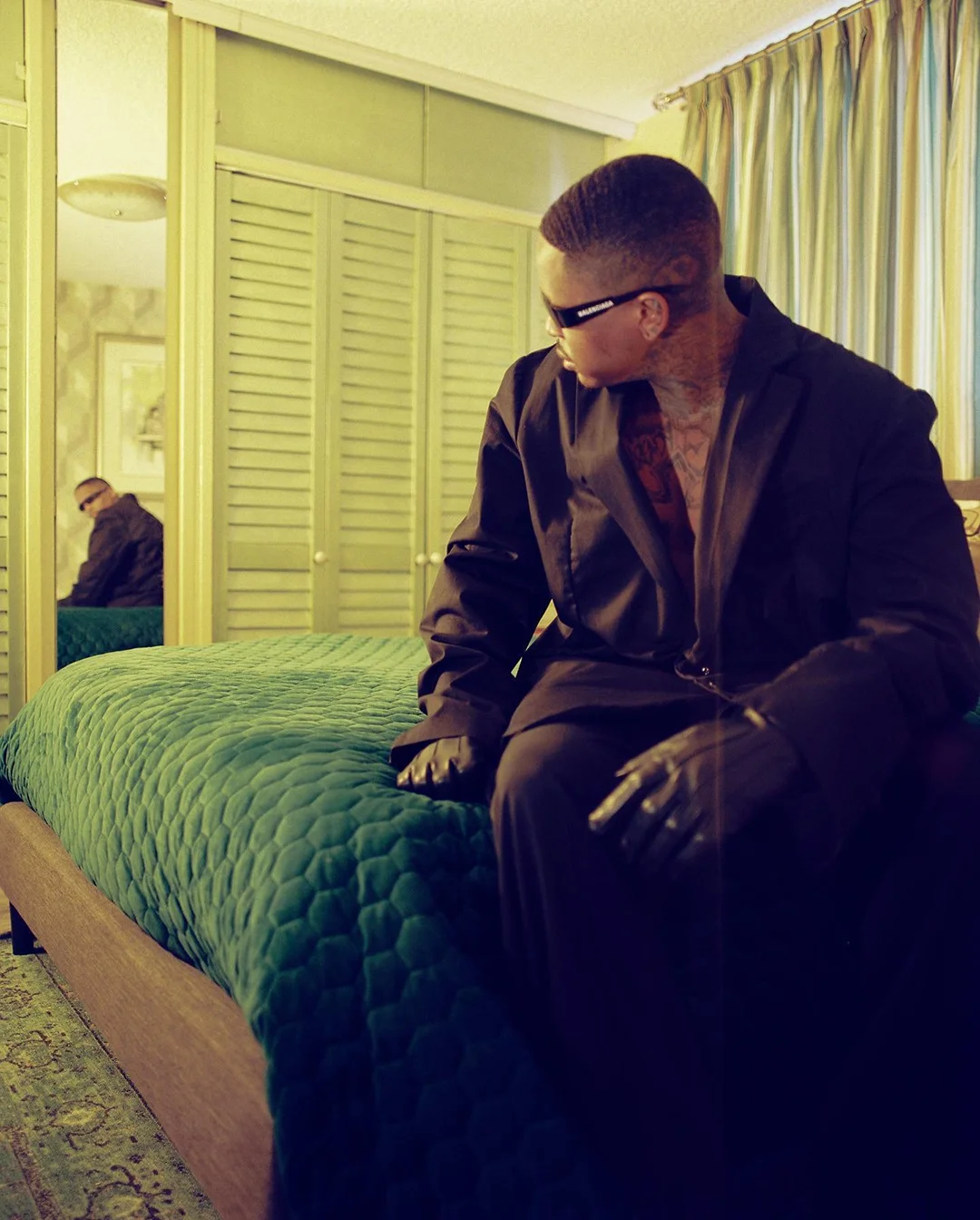 Adult man in a suit sitting on a green quilted bed in a room with a mirror reflecting the subject, creating a surreal, stylish portrait.