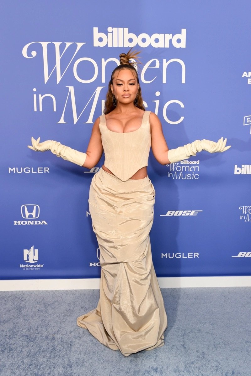 Singer poses on red carpet at Billboard Women in Music event wearing corset top and gown, long gloves, with a billboard backdrop.
