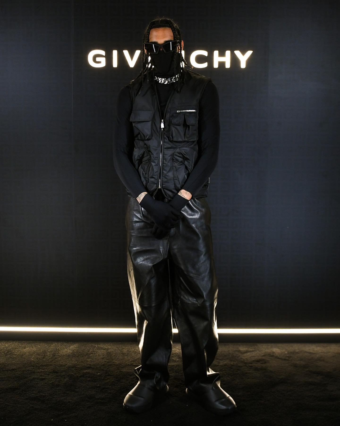 Full-length portrait of a single adult with sunglasses, face covering, vest, and leather pants, standing against a black backdrop with a brand logo.
