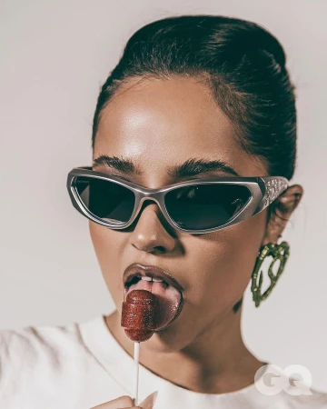 Close-up portrait of a woman in sunglasses licking a lollipop, set against a plain backdrop with studio lighting, featuring a GQ logo.