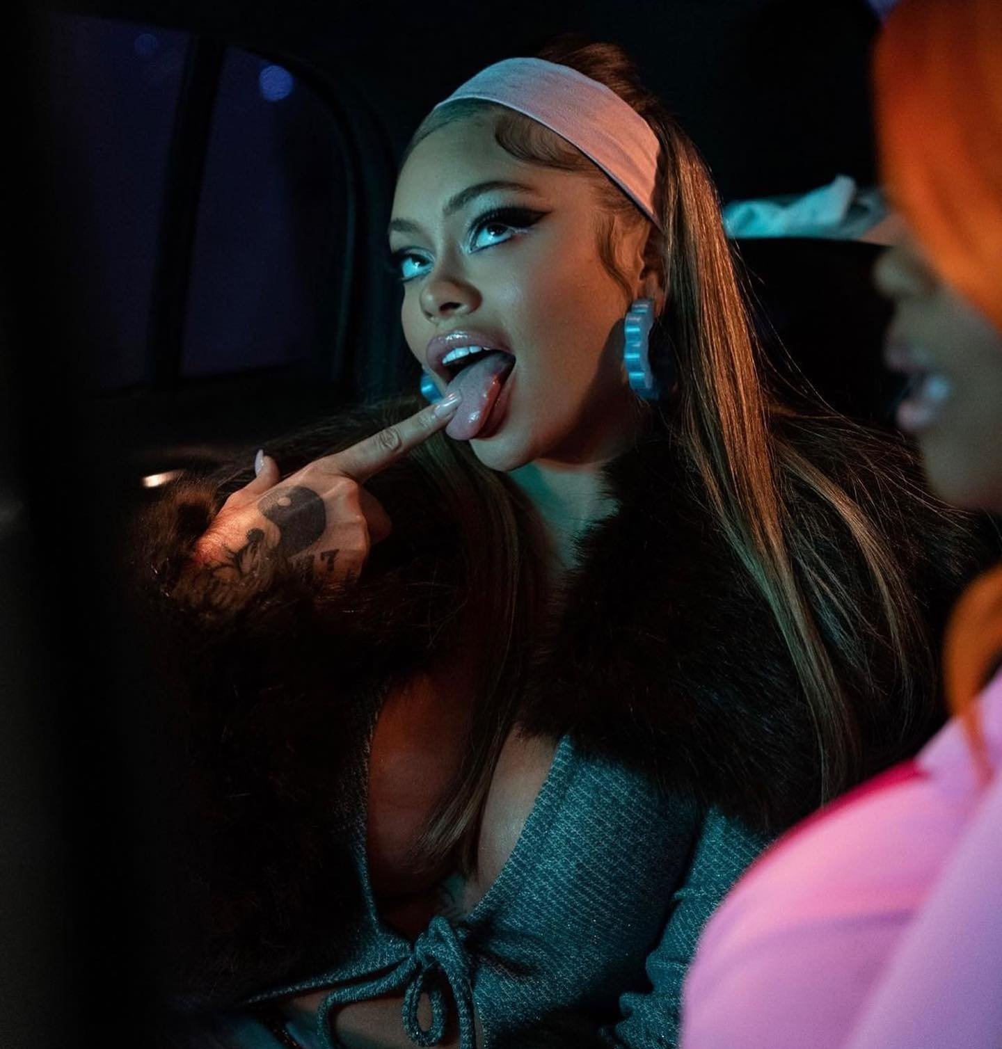 Stylish woman poses with tongue out and finger up, wearing a gray dress, fur coat, headband, and large blue hoop earrings indoors.