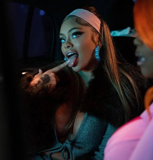 Stylish woman poses with tongue out and finger up, wearing a gray dress, fur coat, headband, and large blue hoop earrings indoors.