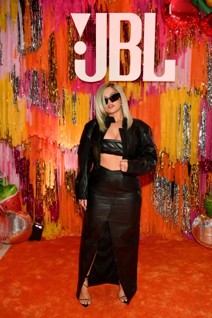 Bebe Rexha poses on orange carpet wearing a black leather outfit, complemented by a vibrant tinsel background and the 'JBL' logo.