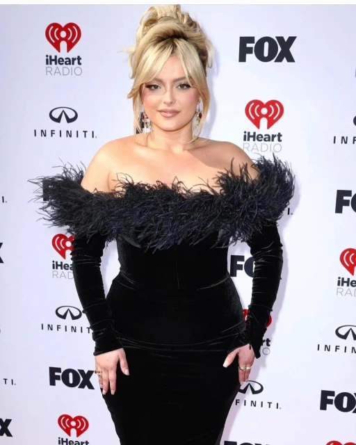 Bebe Rexha poses on a red carpet in a black velvet dress with feather trim, blonde hair styled up, at an indoor entertainment event.
