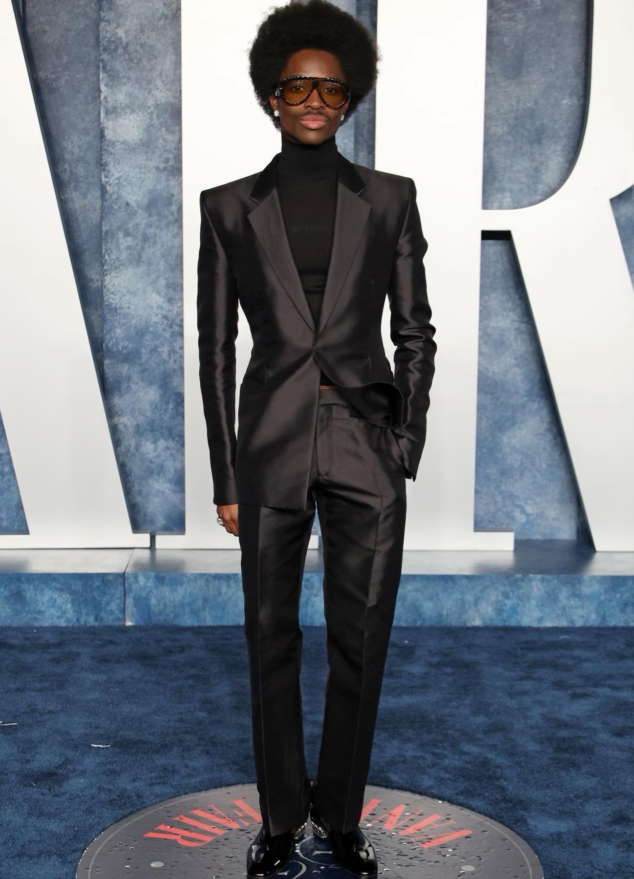 A person in a sleek black suit and sunglasses poses in front of a textured blue and white backdrop, looking directly at the camera.