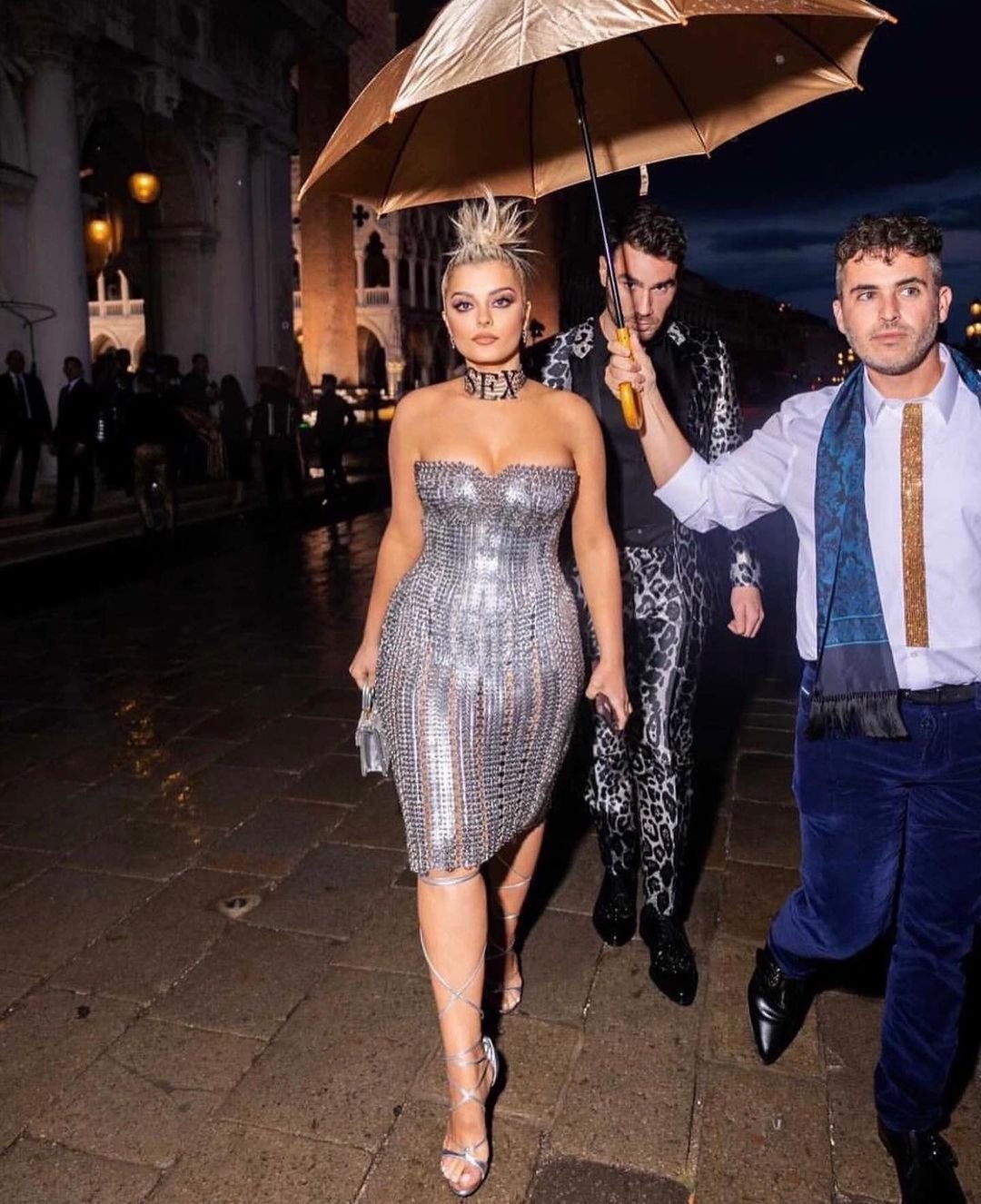 Bebe Rexha poses in a silver dress with two men under an umbrella in Venice. The woman has a choker necklace, and the group is outdoors.