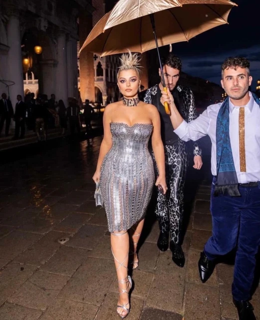 Bebe Rexha poses in a silver dress with two men under an umbrella in Venice. The woman has a choker necklace, and the group is outdoors.