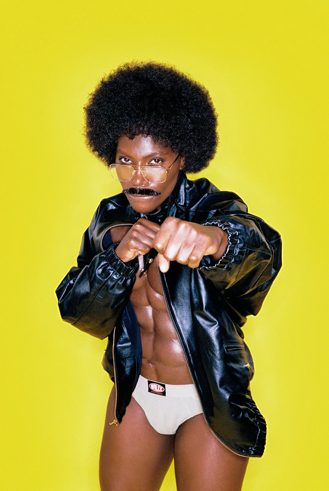 Person in white briefs and leather jacket, flexing abdominal muscles and fists against bright yellow backdrop, wearing glasses and mustache.