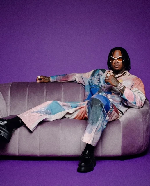 An adult male with sunglasses lounges on a mauve couch against a solid purple backdrop, showcasing patterned matching separates.