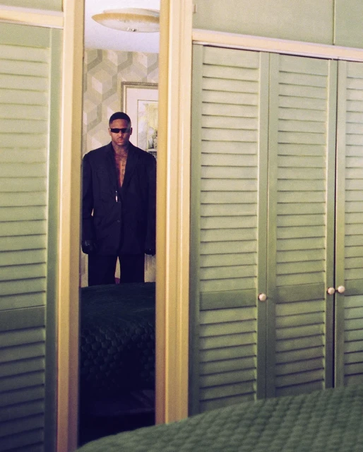 A man in a black suit stands in a doorway reflecting in a mirror with a light green closet in the background, inside an interior.