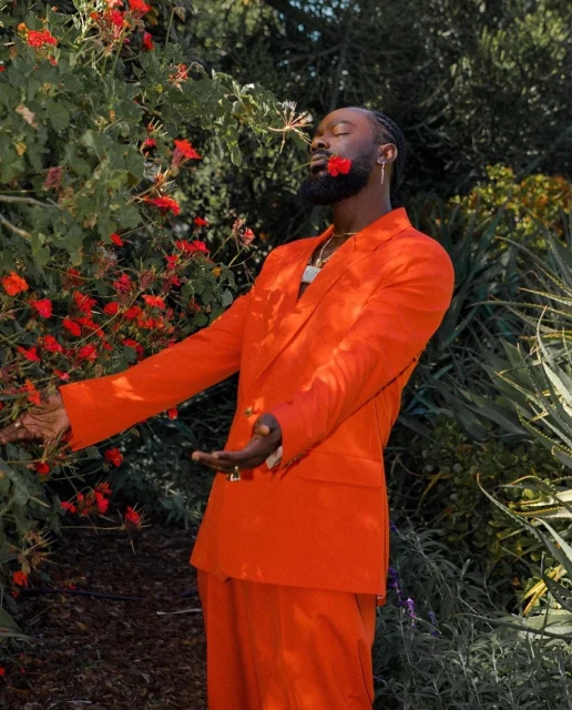 A man in a vibrant orange suit stands outdoors among foliage, a red flower held in his mouth, creating a striking, colorful portrait.