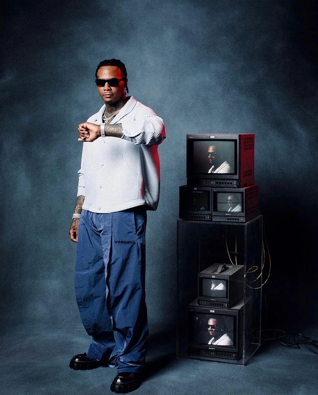 An adult male stands indoors next to stacked television sets while wearing sunglasses, a collared shirt, slacks, and dress shoes.