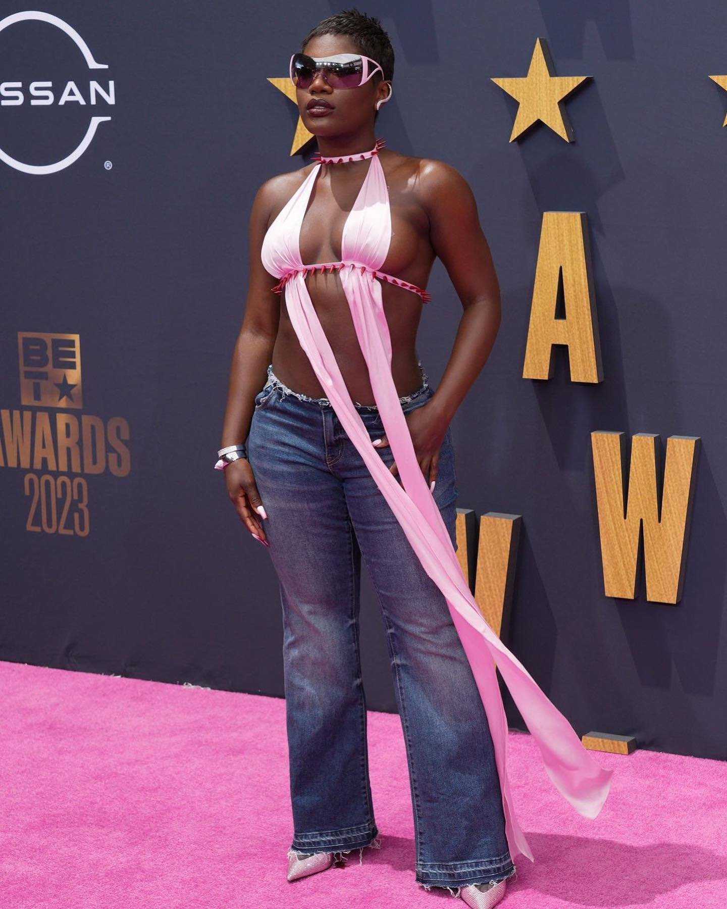 Dark-skinned woman in jeans and pink halter top poses on pink carpet at BET Awards 2023, wearing sunglasses, with Nissan logo in background.