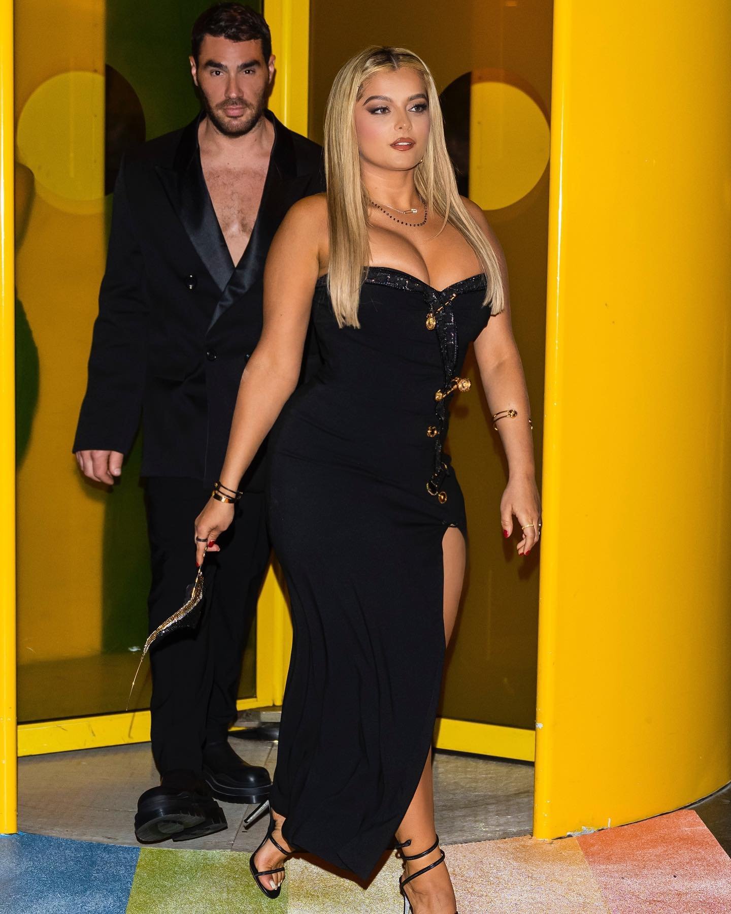 Blonde woman in black dress poses with male companion in suit, accessorized with jewelry and heels, standing indoors near yellow column.