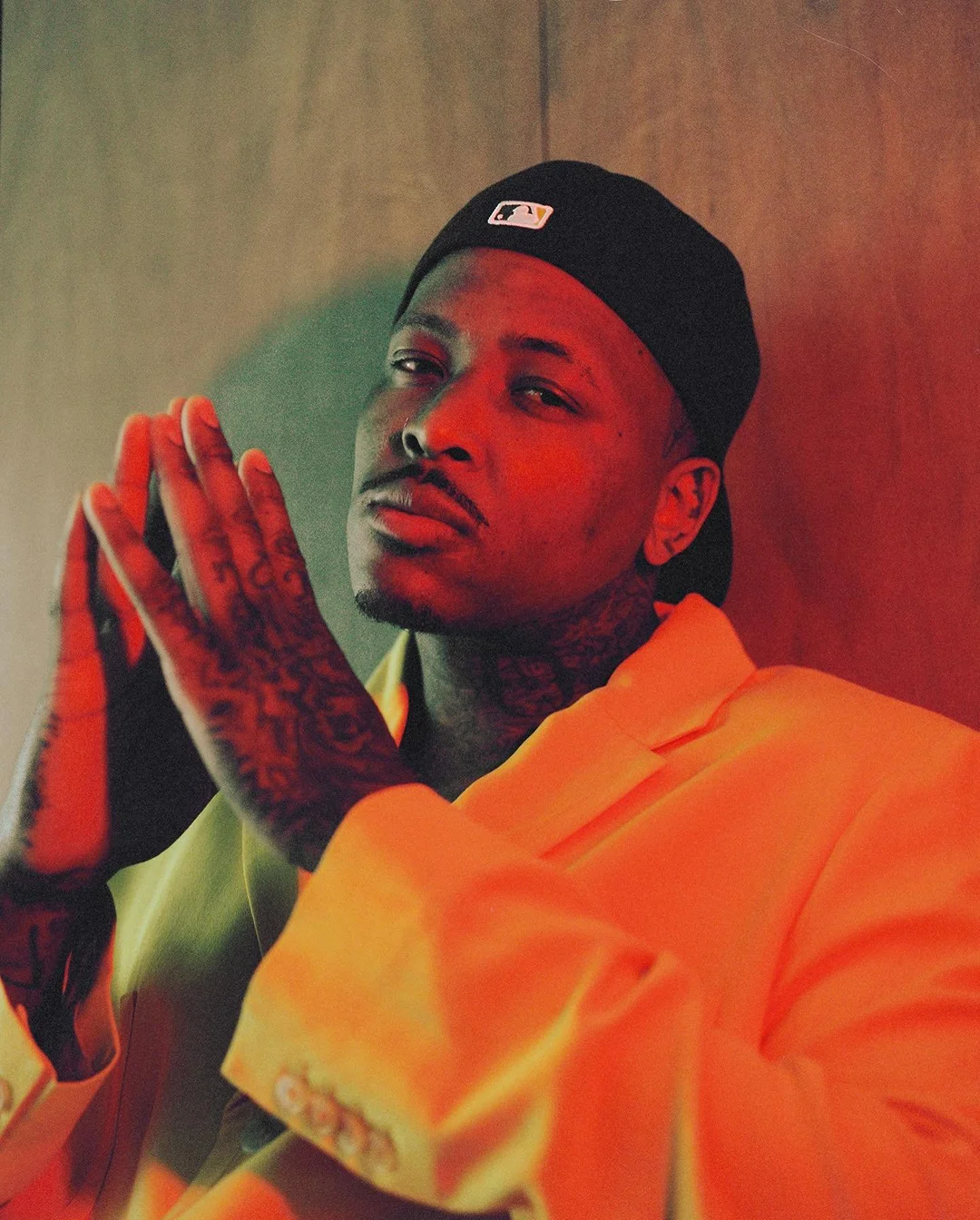 Adult male in a yellow blazer and black cap poses in a studio with hands clasped together, tattoos visible on hands and neck.