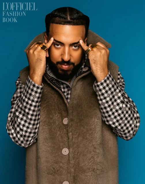 Portrait of French Montana wearing a checked shirt and vest against a blue backdrop, hands near his temples, large gold rings.