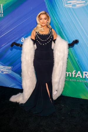 Bebe Rexha poses on a red carpet wearing a black mermaid gown with a faux fur stole and black gloves at an event with sponsor logos.