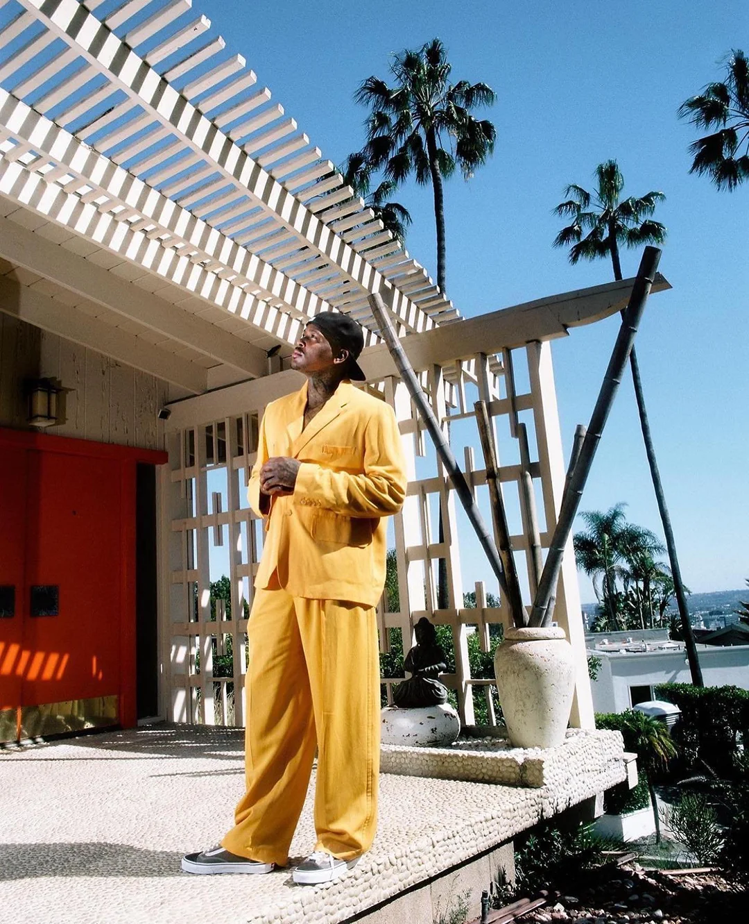 Man in yellow suit poses on a patio with palm trees and mid-century modern home details against a clear blue sky. Relaxed, fashionable moment.