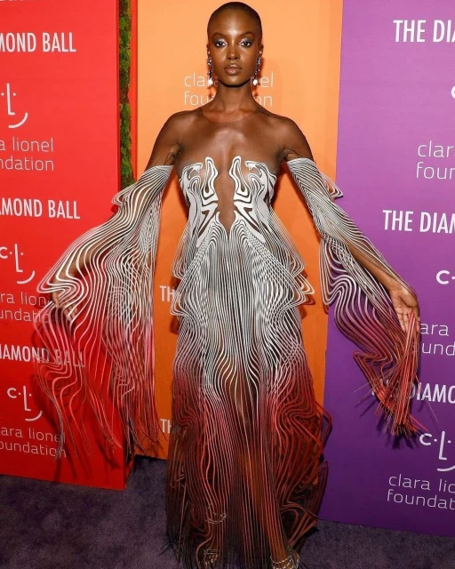 A solo model stands on a red carpet, wearing a striking, semi-transparent, gown with flowing white and red lines and statement earrings.