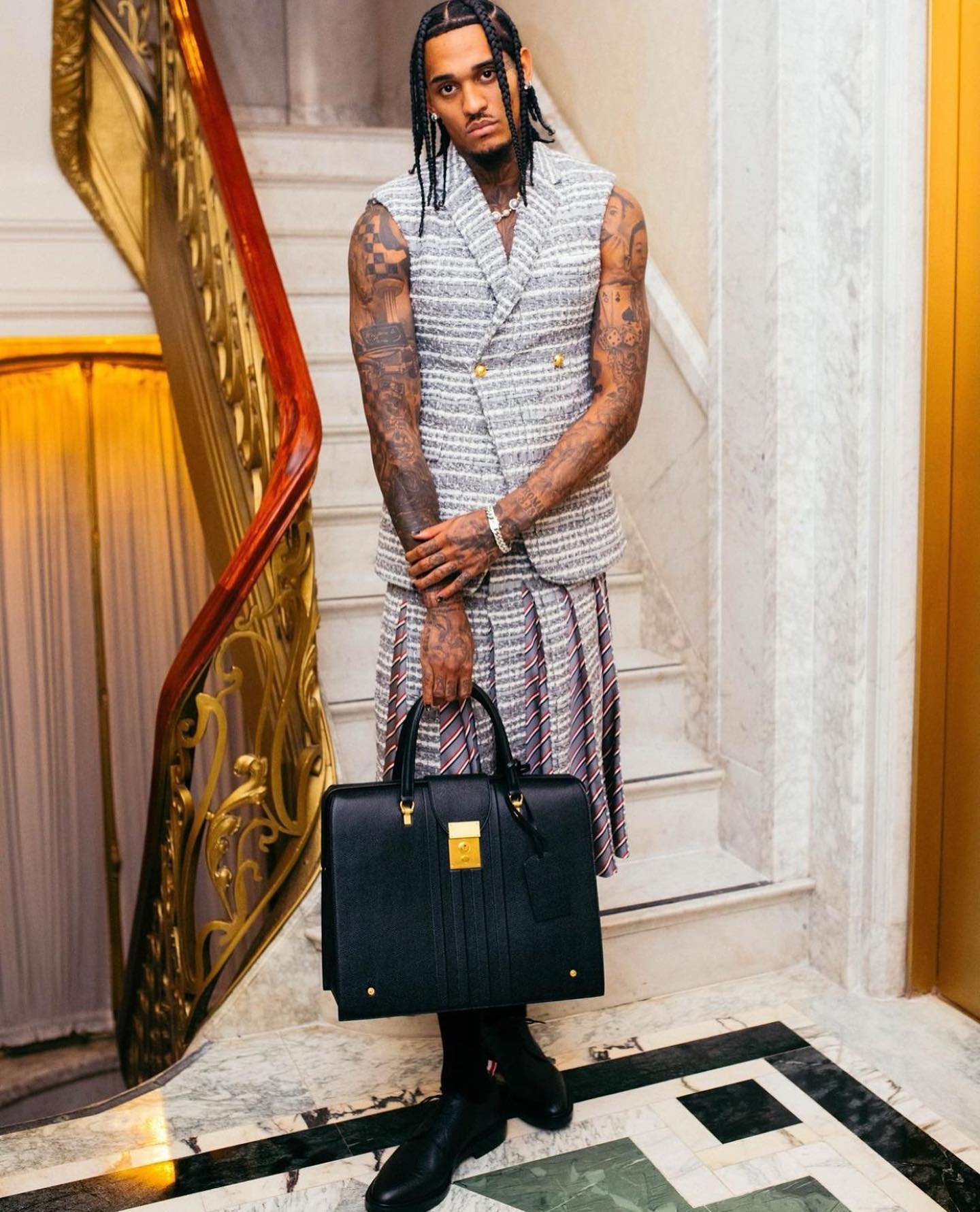 Well-dressed man with tattoos poses indoors wearing a tweed vest, pleated skirt, and carrying a structured black leather briefcase.