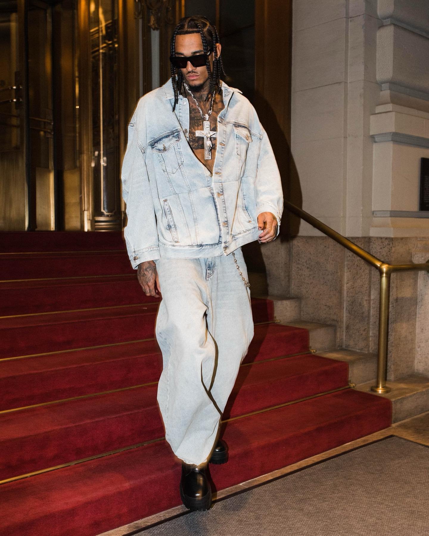 Man in oversized denim jacket and light blue jeans descends red carpeted stairs with gold railing next to a light stone building.