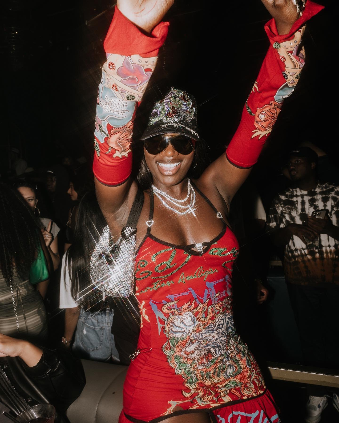Dark-skinned woman in a red patterned dress with matching sleeves dances in a nightclub, wearing sunglasses and jewelry.