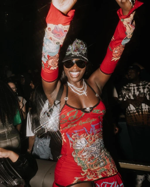 Dark-skinned woman in a red patterned dress with matching sleeves dances in a nightclub, wearing sunglasses and jewelry.