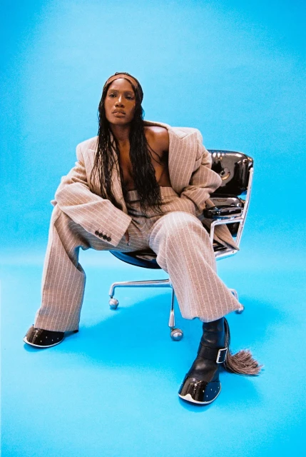 Adult female in a pinstripe suit and patent leather boots sitting in a black office chair against a cyan background in a studio shot.