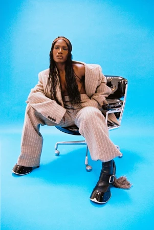 Adult female in a pinstripe suit and patent leather boots sitting in a black office chair against a cyan background in a studio shot.