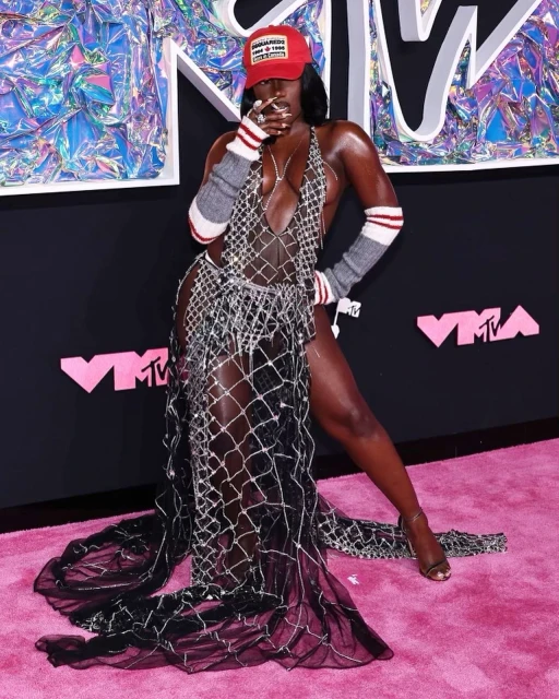 Woman on a pink carpet wearing a sparkly dress, red cap, and grey sleeves at the MTV Video Music Awards (VMAs).