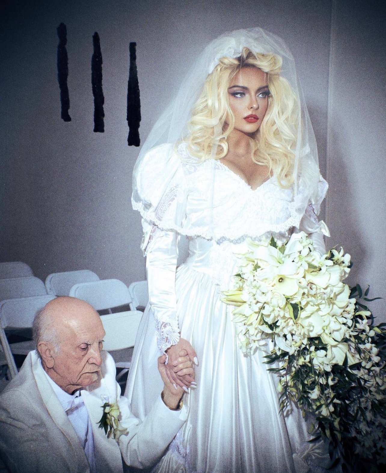 An elderly man in a suit holds the hand of a young woman in a white wedding dress, with a bouquet, in an indoor setting.