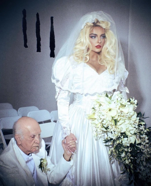 An elderly man in a suit holds the hand of a young woman in a white wedding dress, with a bouquet, in an indoor setting.
