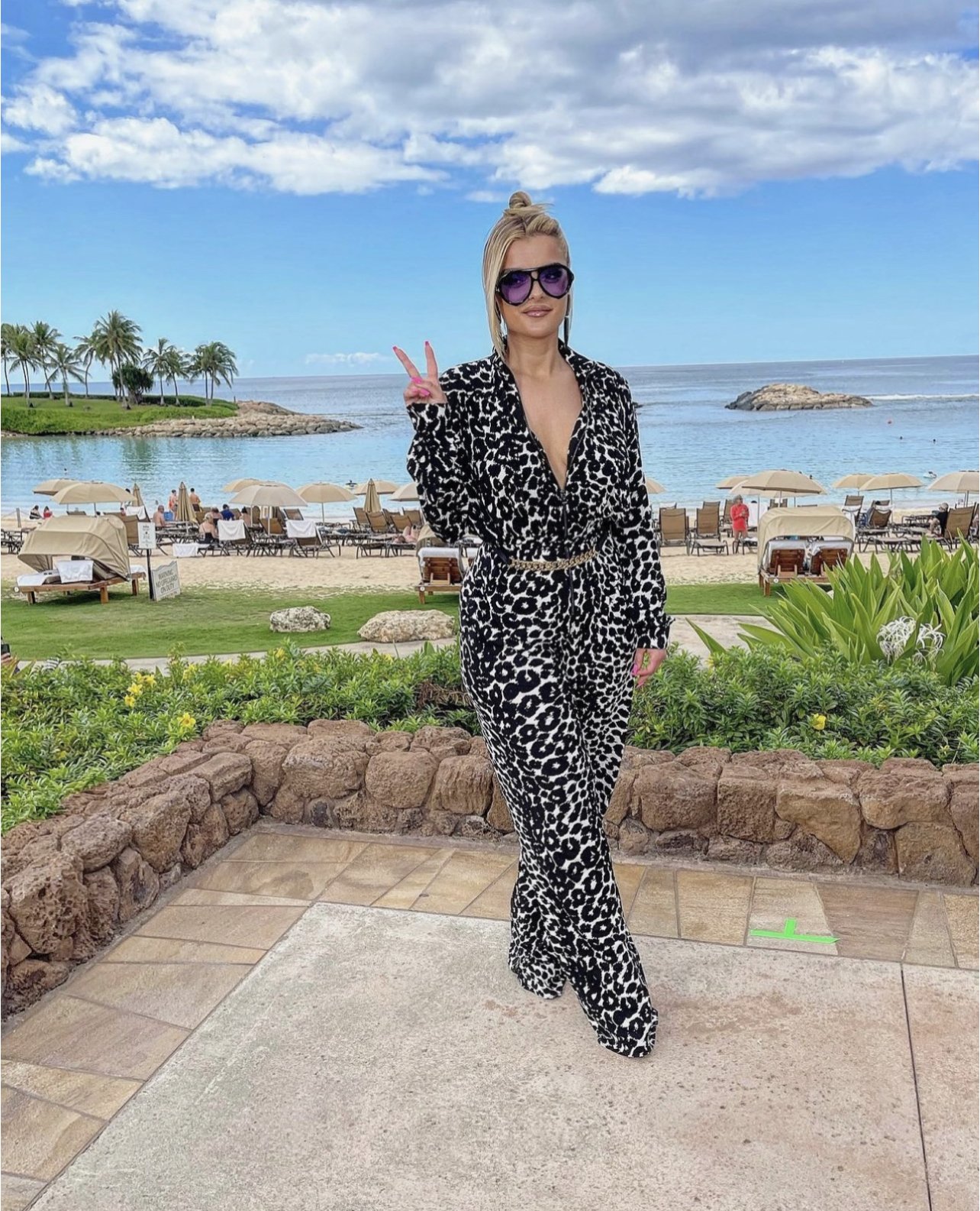 Adult female wearing a patterned jumpsuit and sunglasses standing on a stone patio with a beach and ocean in the background.