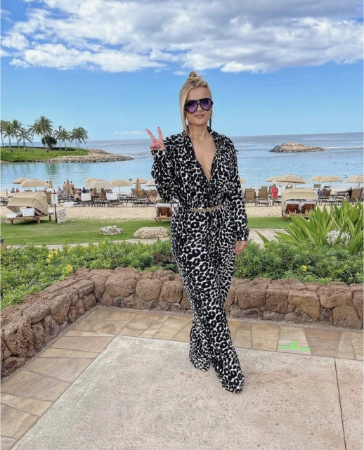 Adult female wearing a patterned jumpsuit and sunglasses standing on a stone patio with a beach and ocean in the background.