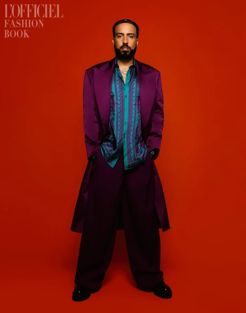Portrait of a bearded man in a purple suit, patterned shirt, and black shoes, standing against a vibrant orange background.