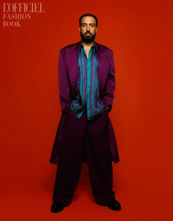 Portrait of a bearded man in a purple suit, patterned shirt, and black shoes, standing against a vibrant orange background.
