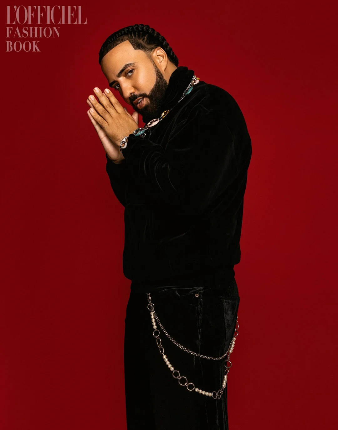 Portrait of French Montana against a red background. He is wearing a black velvet jacket, jewelry, and black pants with pearl chain.