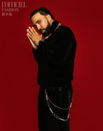 Portrait of French Montana against a red background. He is wearing a black velvet jacket, jewelry, and black pants with pearl chain.