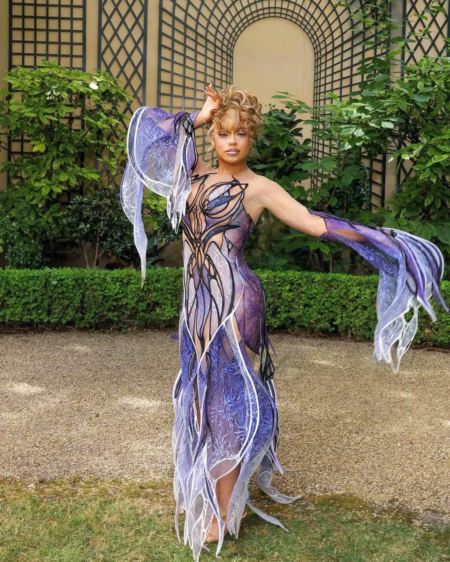Woman wearing a custom purple and white gown poses in a garden with trellises and greenery in the background, under natural lighting.