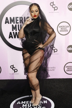 Adult female poses on a red carpet in a black bodysuit with sheer panels and a black veil at the American Music Awards.