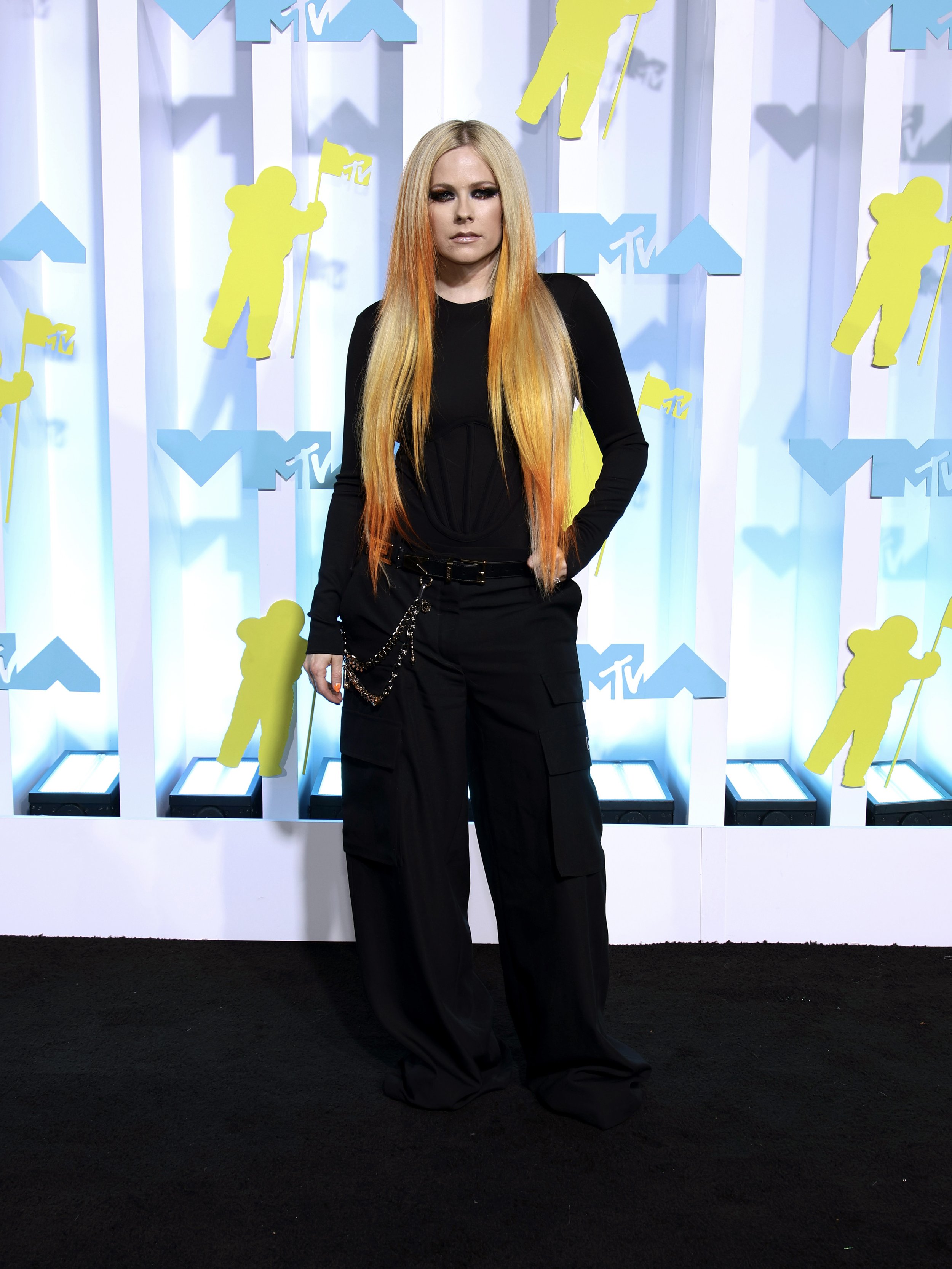 Avril Lavigne poses on the red carpet at an event with a printed backdrop, wearing black pants and a long-sleeve black top.
