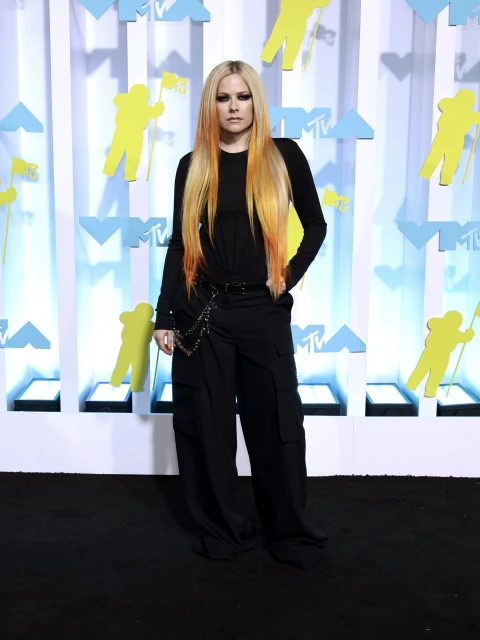 Avril Lavigne poses on the red carpet at an event with a printed backdrop, wearing black pants and a long-sleeve black top. 
