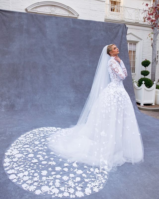 Woman in white long-sleeved wedding dress with long train and veil poses in front of gray backdrop and building exterior.