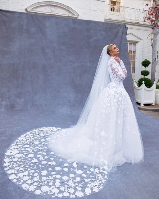 Woman in white long-sleeved wedding dress with long train and veil poses in front of gray backdrop and building exterior.