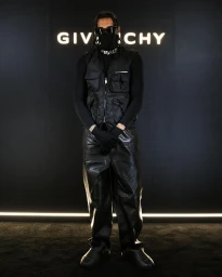 Man in all-black streetwear outfit wearing sunglasses and face mask standing before a backlit Givenchy sign