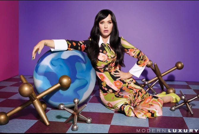 Katy Perry poses in a colorful floral dress with large jacks toys on a checkered floor against a violet and pink studio backdrop.