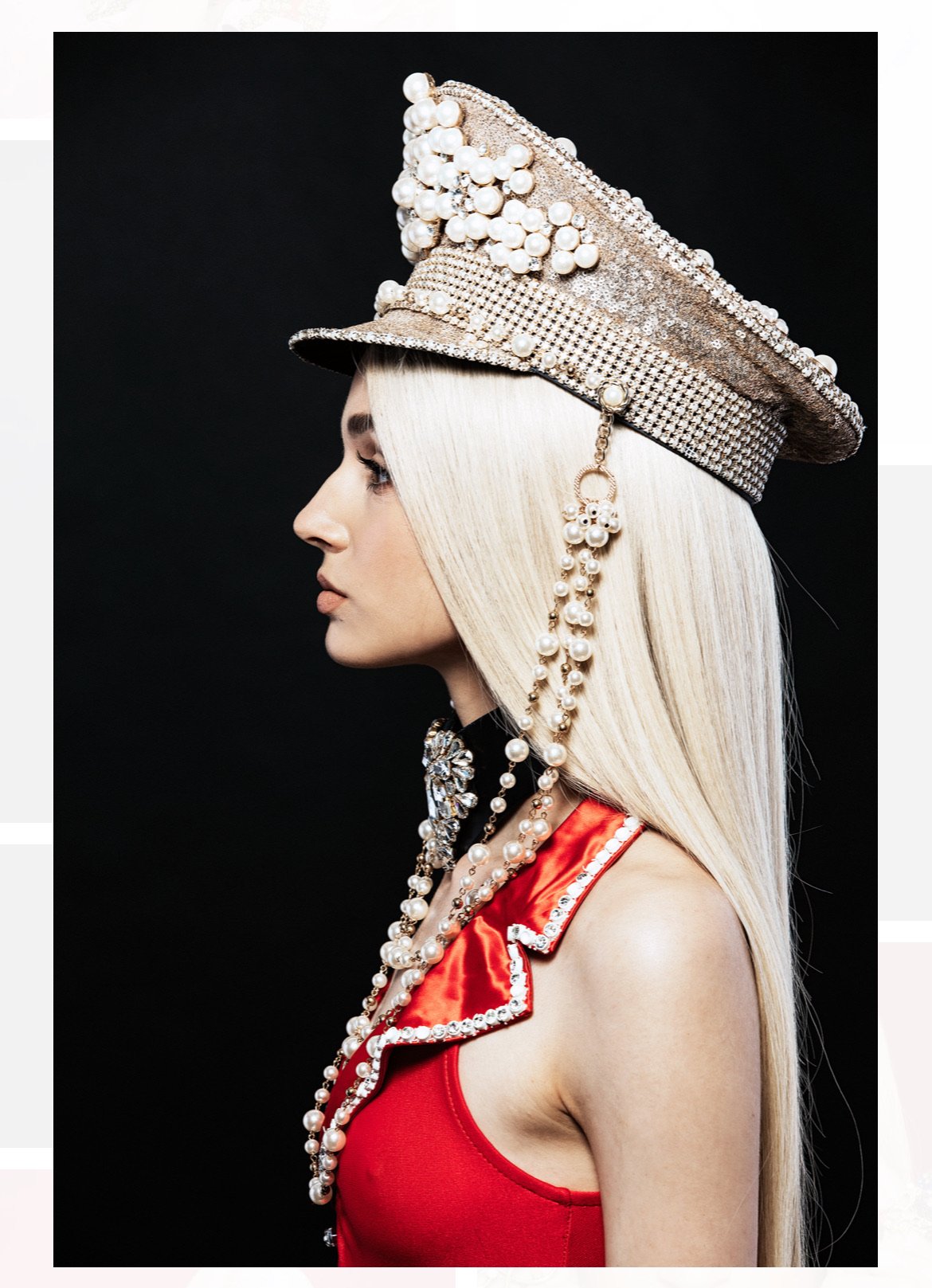 Side profile of an adult female with platinum blonde hair, wearing a red top, pearl jewelry, and a beaded military-style cap against a black background.