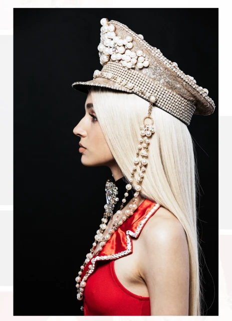 Side profile of an adult female with platinum blonde hair, wearing a red top, pearl jewelry, and a beaded military-style cap against a black background.